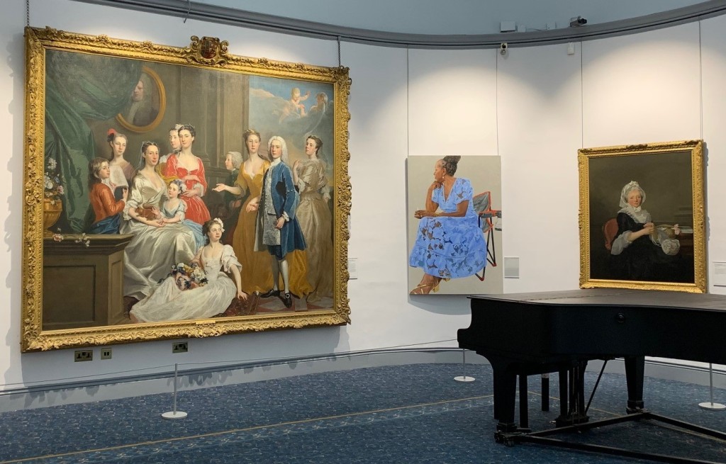 Installation view of three fine art portraits displayed on the walls at Wolverhampton Art Gallery near to a grand piano. The central portrait depicts "Ann" (2022) - a contemporary representation of the artist's sister, seated on a foldaway chair.